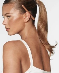 Profile of a woman with a sleek high ponytail and glowing skin, wearing a white tank top with a nude cosmetic pencil behind her ear.