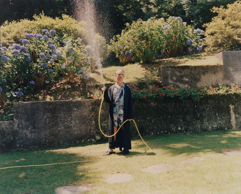 Man standing on a lawn wearing a dark robe and floral set, holding a yellow garden hose spraying water upwards in a sunny garden.