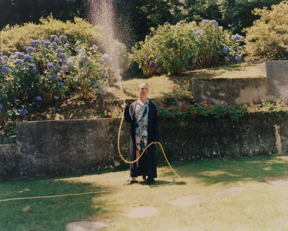 Man standing on a lawn wearing a dark robe and floral set, holding a yellow garden hose spraying water upwards in a sunny garden.