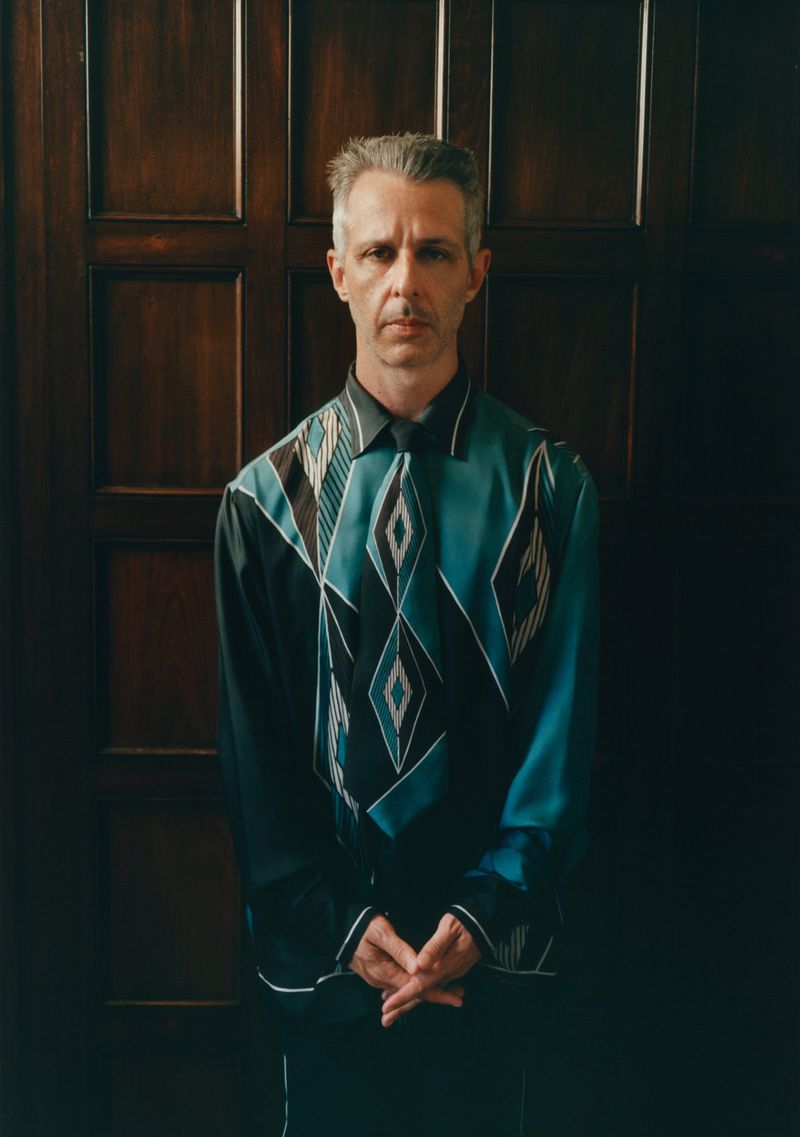 A man with short grey hair wearing a black and teal geometric patterned shirt standing against a dark wooden paneled door.