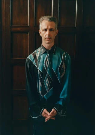 A man with short grey hair wearing a black and teal geometric patterned shirt standing against a dark wooden paneled door.