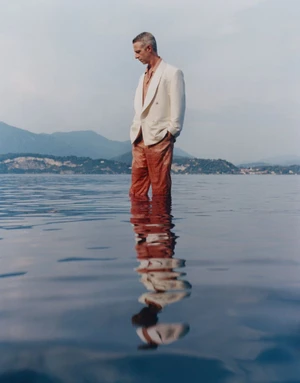 Actor Jeremy Strong stands submerged in calm lake water wearing an ivory blazer and terracotta trousers, looking downward.