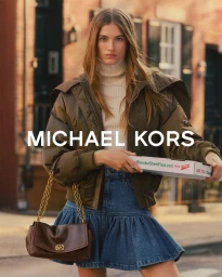 Model carrying a brown chain-strap handbag and holding a pizza box on a city street wearing an olive jacket and denim skirt.