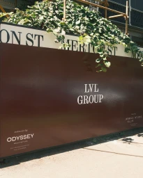 Branded dark brown construction hoarding for LVL Group with typography and green foliage draped along the top edge.