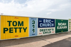 Painted shipping container wall displaying musical artist names including Tom Petty, Eric Church, and Noah Kahan outdoors.