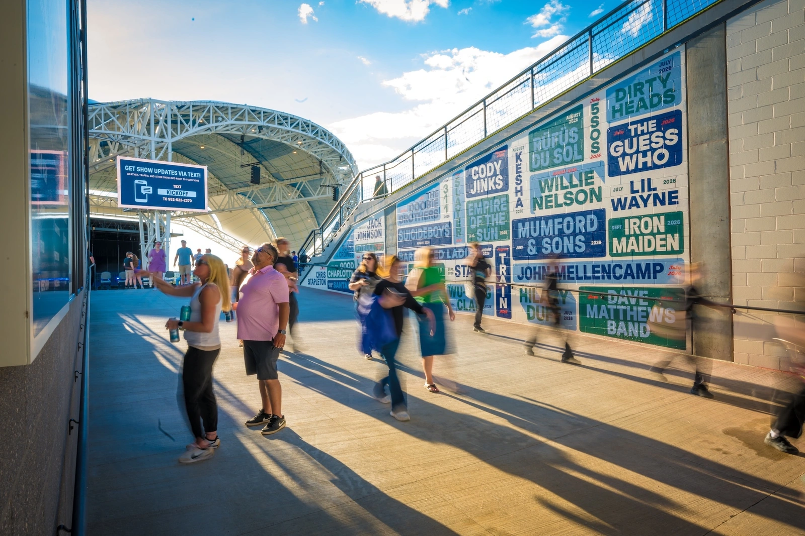 Concert attendees walk along an outdoor amphitheater concourse with a large typographic artist mural wall under a sunny sky.
