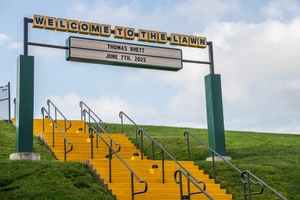 Yellow outdoor staircase leading up a grassy hill beneath a Welcome to the Lawn marquee sign under a partly cloudy sky.