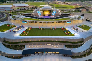 High-angle aerial view of the outdoor Mystic Lake Amphitheater with an arched concert stage, tiered lawn, and box office entrance.