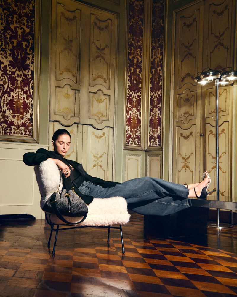 A woman reclining in a white shearling chair, holding a black handbag, feet resting on a table, inside a vintage-style room.
