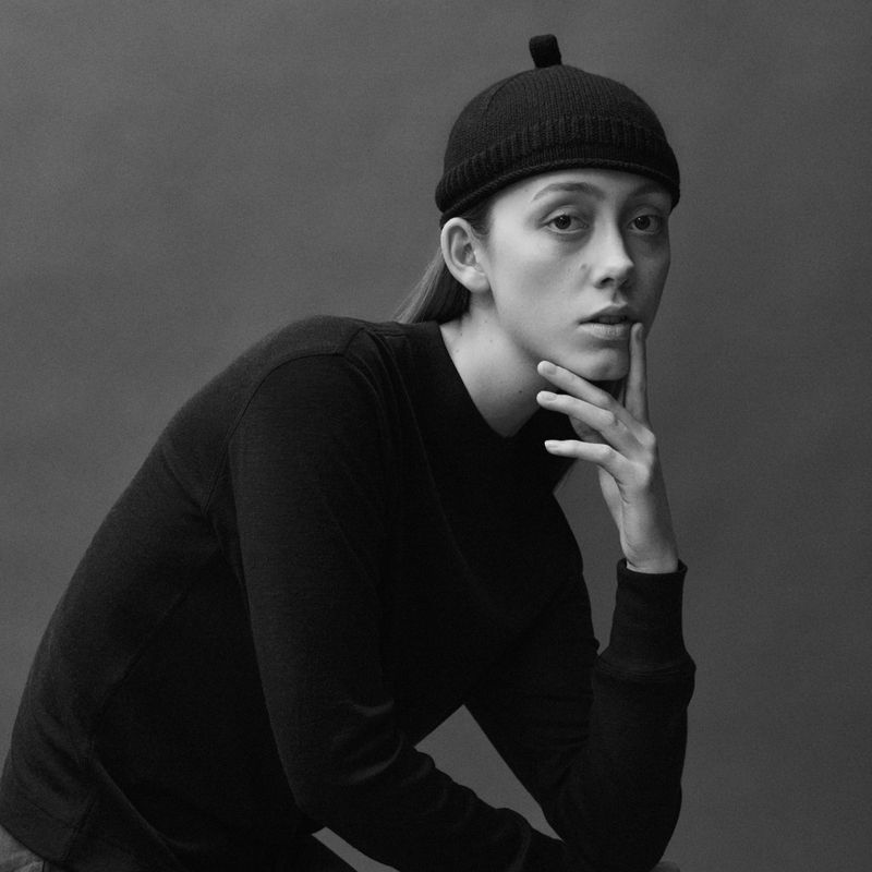 Portrait of a woman wearing a black knit beanie and long-sleeved sweater, posing with her hand resting on her chin against a dark background