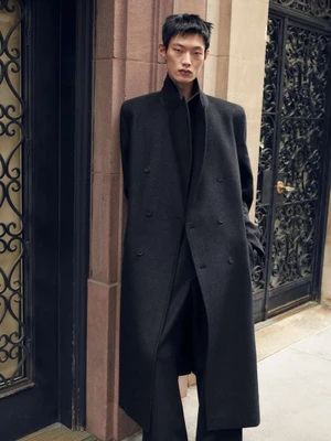 East Asian male model wearing a long black tailored wool overcoat standing against a stone doorway with ornate wrought iron gates.