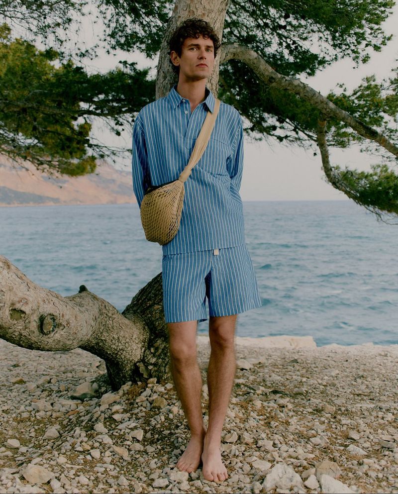 Young man poses under tree in striped shirt and shorts near water. A woven bag is slung over his shoulder in an outdoor setting.