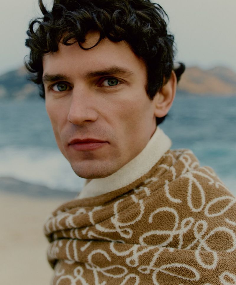 Close-up portrait of a man with curly hair, blue eyes, wrapped in a patterned brown and white towel against an ocean backdrop.