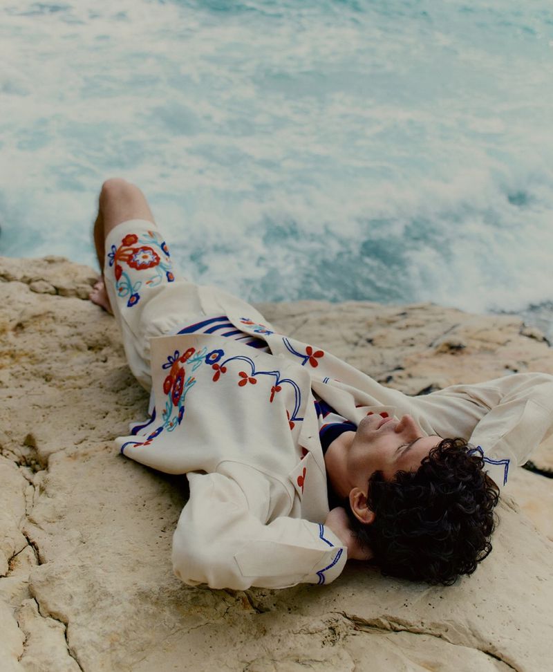 Relaxed adult male with dark curly hair lies on a rock formation near a turquoise ocean. He is wearing an embroidered casual outfit.