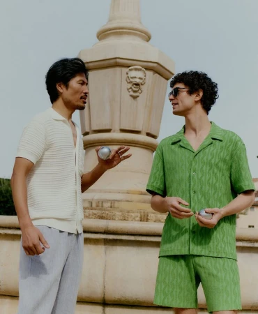 Two men in casual summer attire stand outdoors near a stone fountain, holding boules in their hands, with a light sky background.