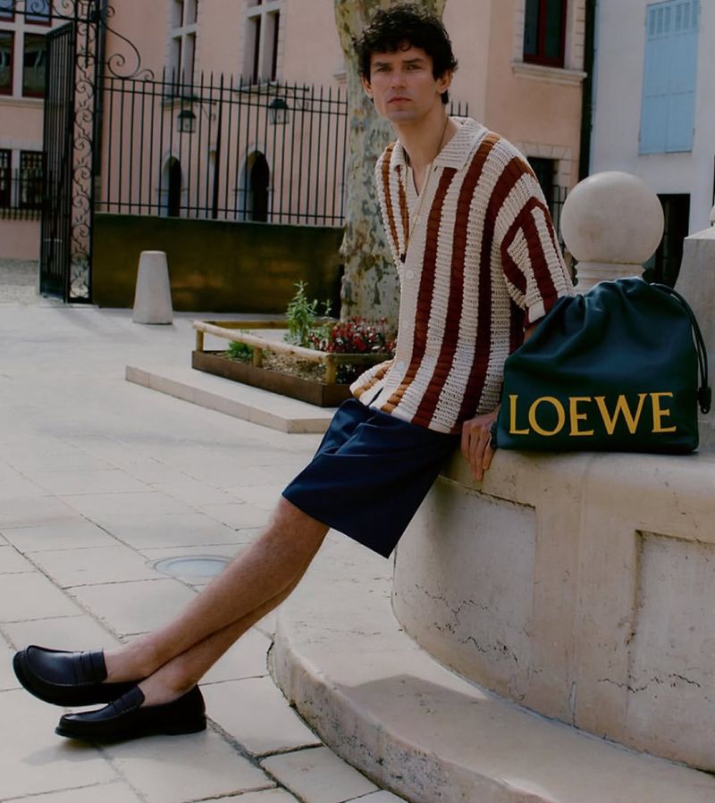 A young adult man with dark hair sits outside on a ledge with a designer Loewe bag. He wears a striped shirt and shorts.