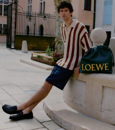 A young adult man with dark hair sits outside on a ledge with a designer Loewe bag. He wears a striped shirt and shorts.