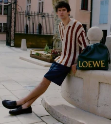 Model Arthur Gosse in a striped knit polo and navy shorts seated outdoors on a stone fountain ledge beside a dark green Loewe bag.