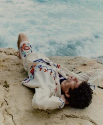 Arthur Gosse reclining on rocky coastal stone by churning ocean water wearing embroidered cream jacket and shorts