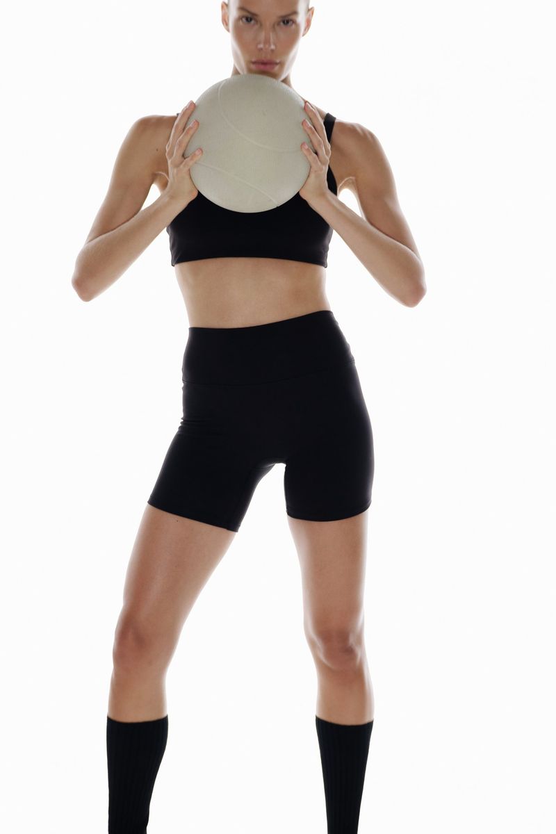 Model with shaved head wearing black athletic crop top and shorts holding a light grey volleyball against a white background.
