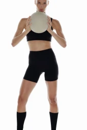 Model Alena Blohm posed in a black sports bra and biker shorts holding a white ball against a white studio background.