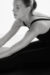 Monochrome shot of Alena Blohm in black activewear stretching forward while seated on a studio floor against a white backdrop.
