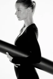 Model Alena Blohm in profile wearing a black long-sleeve top and holding a rolled black fitness mat against a white background.