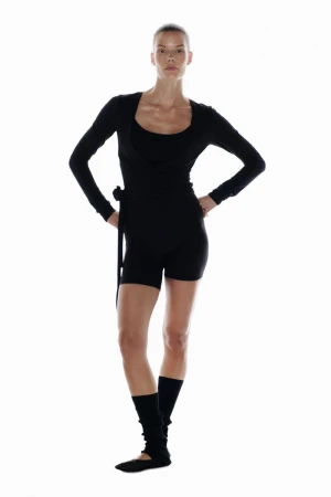 Model Alena Blohm poses in a black long-sleeve wrap top, black shorts, and leg warmers against a solid white studio background.