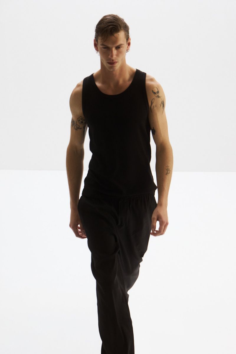 A man with tattoos on his arms wearing a black tank top and black trousers walks towards the camera against a white background.