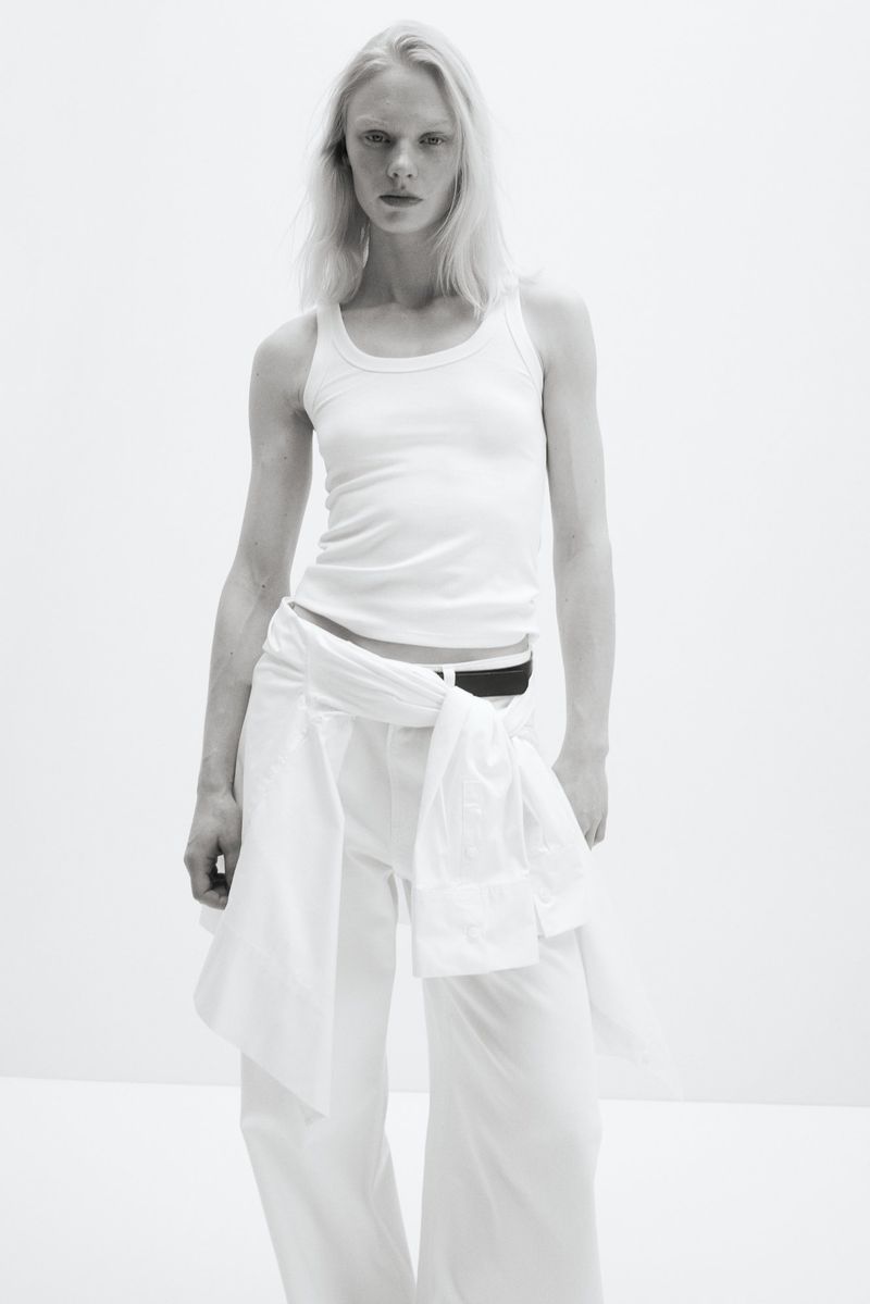 Model Vilma Sjöberg in a white tank top and white trousers with a shirt tied at the waist against a white studio background.