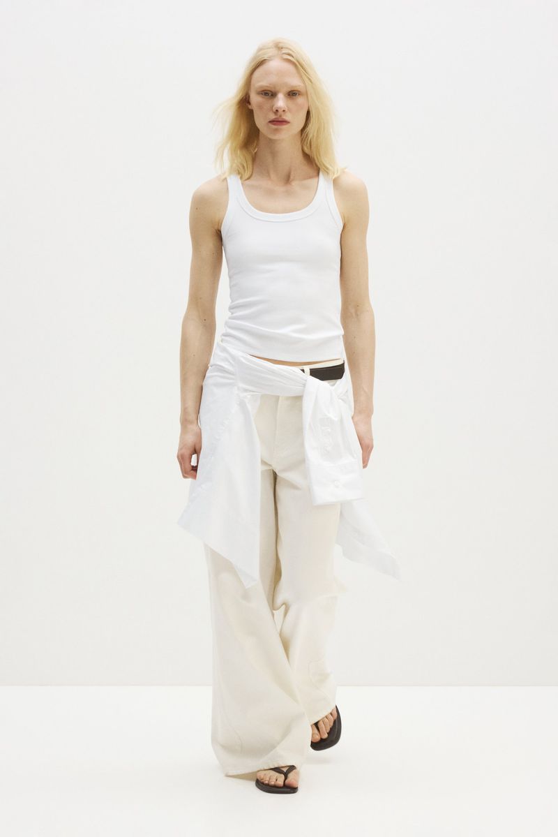 Model wearing a white tank top, cream wide-leg trousers, and a white shirt tied at the waist against a white studio background.