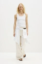 Model wearing a white tank top, cream wide-leg trousers, and a white shirt tied at the waist against a white studio background.