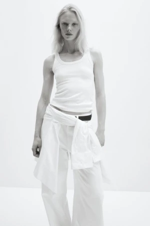 Model Vilma Sjöberg in a white tank top and white trousers with a shirt tied at the waist against a white studio background.