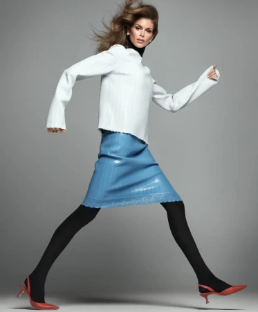 Kaia Gerber posing mid-stride in a white long-sleeve top, blue skirt, black tights, and red slingback heels against a grey backdrop.