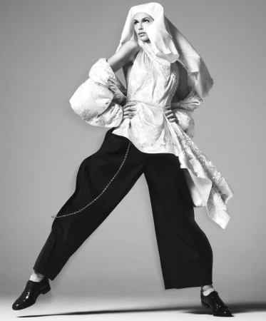 Model Kaia Gerber wearing a white draped headpiece, brocade tunic, and wide black trousers posing dynamically against a grey studio backdrop.