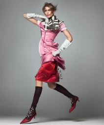 Model Kaia Gerber leaping mid-air wearing a pink satin dress, red embellished skirt, striped scarf, white gloves, and red heels.