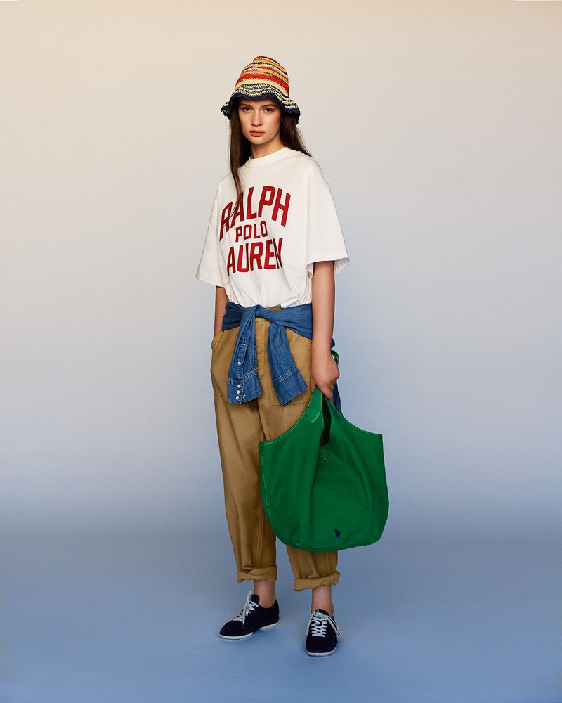 Full-length studio shot of a young female model wearing a white Ralph Lauren t-shirt, khaki pants, and a colorful knit bucket hat, holding a green tote bag.