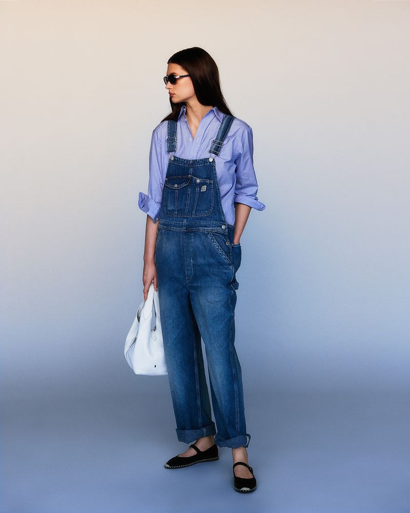 Model wearing blue denim overalls, a light blue collared shirt, sunglasses, and black flats while holding a white tote bag.