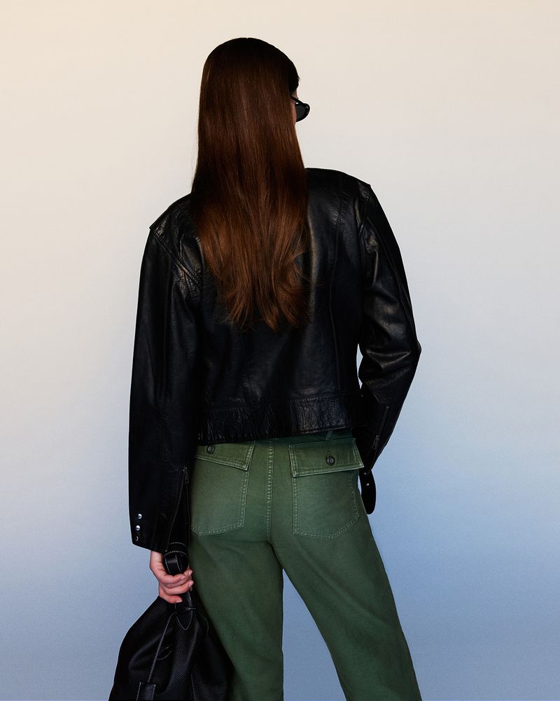 A woman with long brown hair wears a black leather jacket and green pants, standing in front of a plain white background.