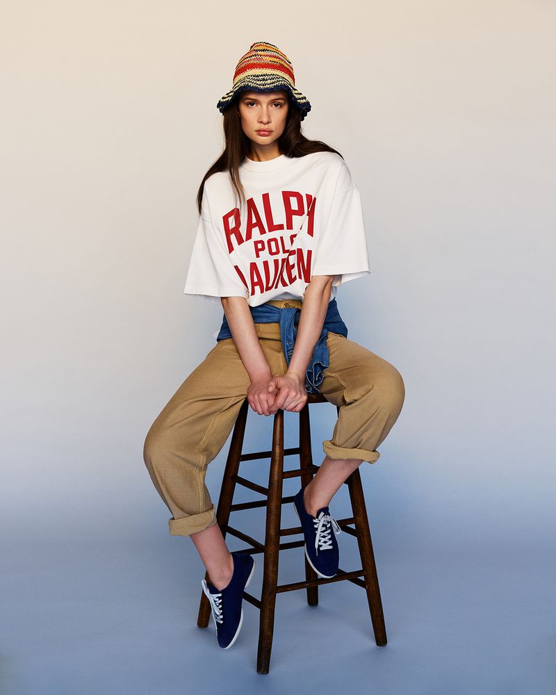 A young woman sits on a wooden stool wearing a Ralph Lauren t-shirt, denim jacket, and striped hat in a studio setting.