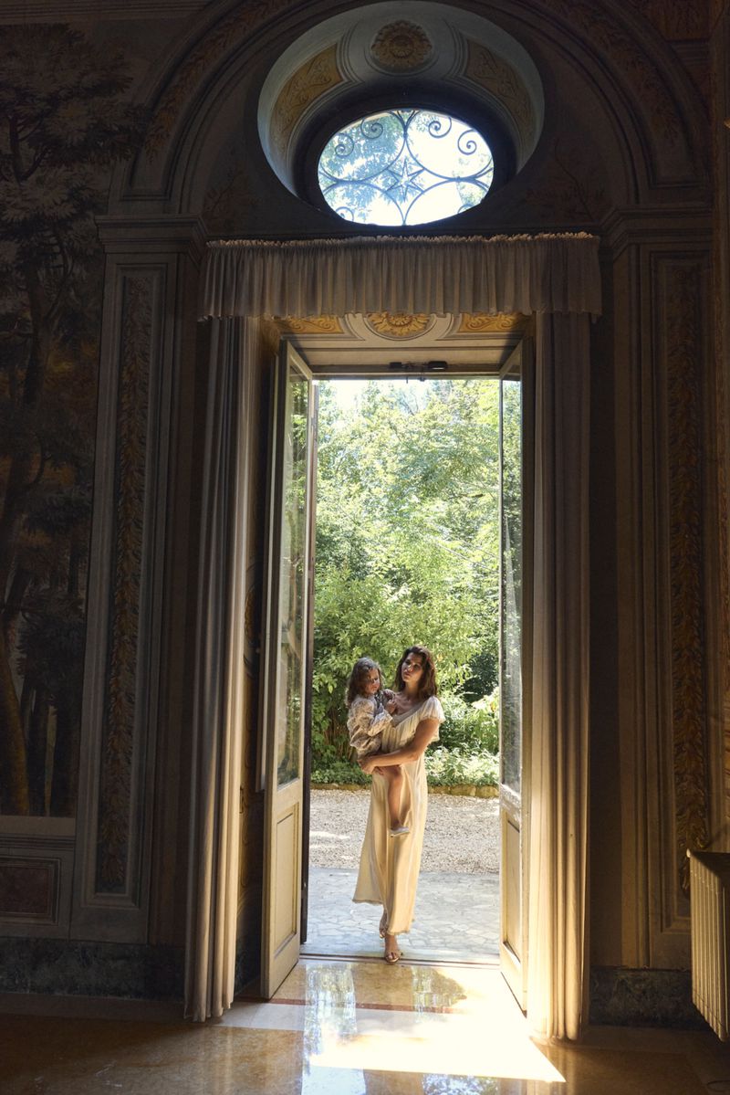 Georgia Fowler holding a child while standing in a tall open doorway overlooking a sunlit garden.