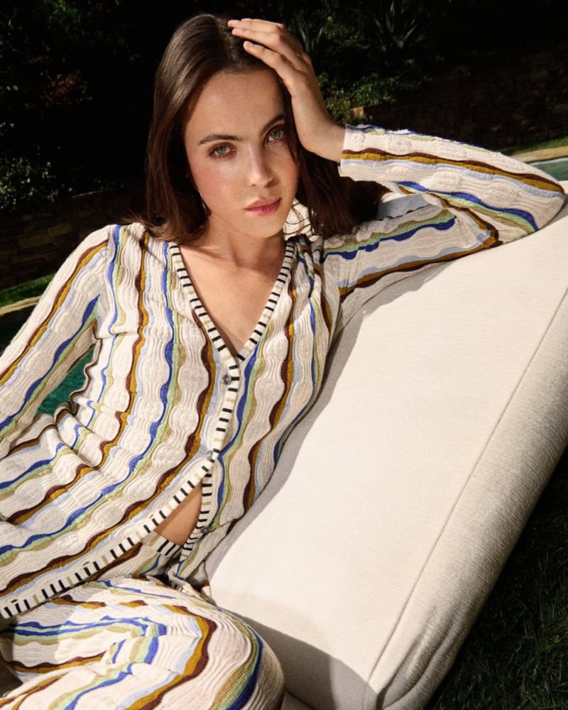 Attractive brunette woman poses outdoors wearing a patterned knit sweater and pants set while relaxing on a white sofa in a garden.