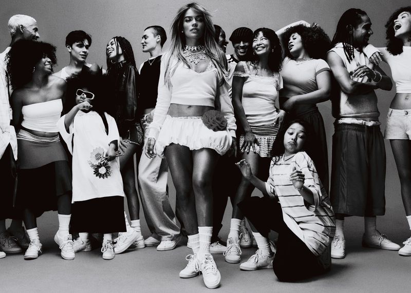 Karol G stands centrally in a black-and-white group portrait, wearing a cropped long-sleeve top and mini skirt, surrounded by diverse people.
