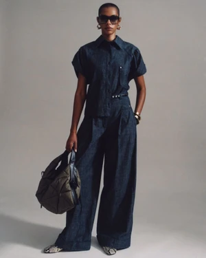 Model wearing dark denim shirt and trousers holding a dark quilted bag against a grey seamless background