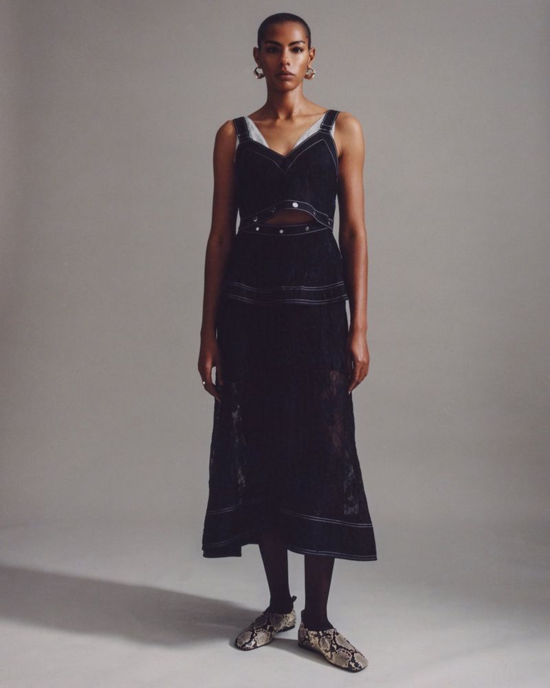 A woman stands in a studio wearing a black sleeveless dress with contrasting white stitching and snake-print flats, posing for a portrait.