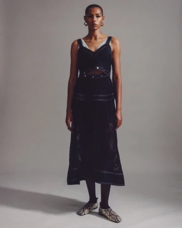 A woman stands in a studio wearing a black sleeveless dress with contrasting white stitching and snake-print flats, posing for a portrait.