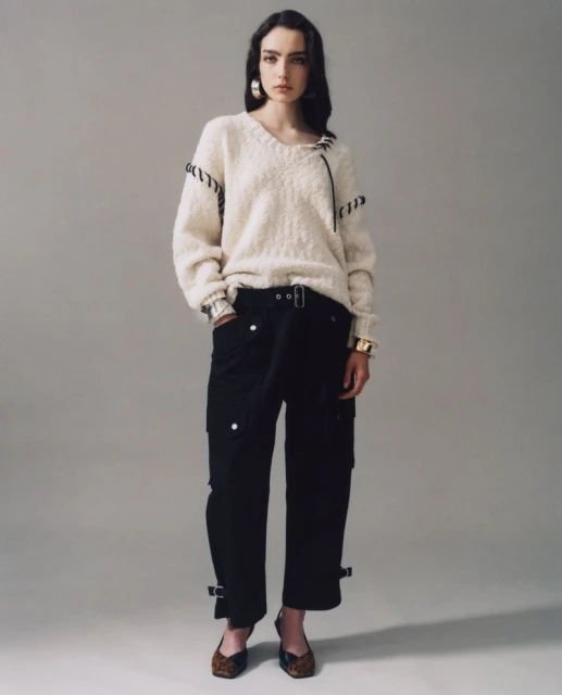 Fashion portrait of a woman wearing a cream-colored sweater and black pants against a neutral backdrop, showcasing modern style.