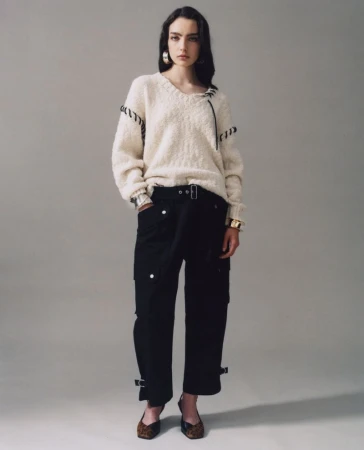 Fashion portrait of a woman wearing a cream-colored sweater and black pants against a neutral backdrop, showcasing modern style.