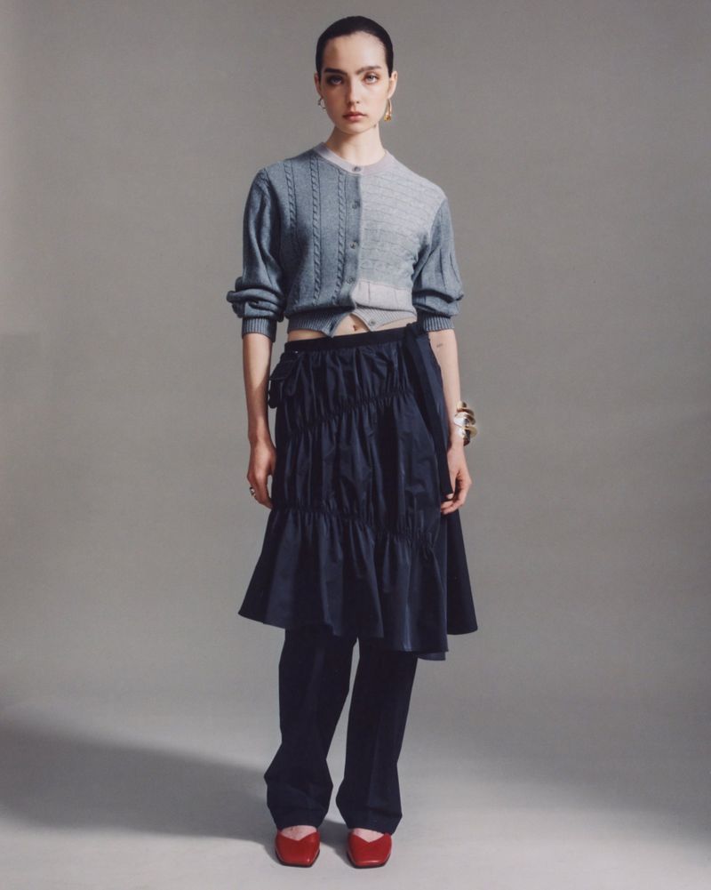 Full-length studio shot of a woman in a two-tone cardigan, layered skirt, pants, and red flats against a neutral grey background.