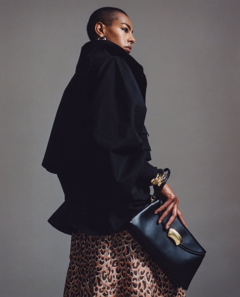 Adult female in a black jacket poses with a black clutch purse and a leopard print skirt against a plain studio backdrop.